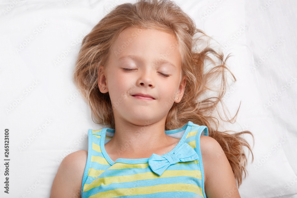Healthy appealing little girl with long messy hair, keeps eyes shut, dressed in pyjamass, has pleasant dreans during night rest, lies on white bed. Small child has healthy sleep. Rest concept Healthy appealing little girl with long messy hair, keeps eyes shut, dressed in pyjamass, has pleasant dreans during night rest, lies on white bed. Small child has healthy sleep. Rest concept
