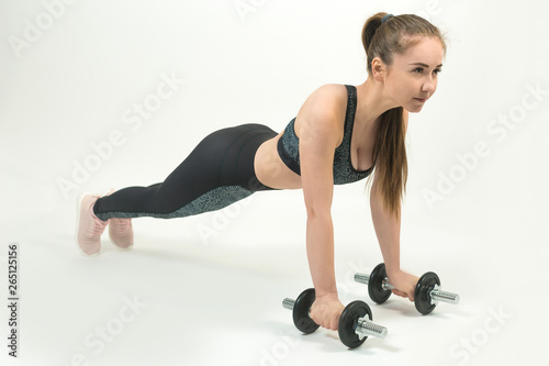Slim fit fitness girl performs a set of exercises on a white background. A girl in sporty tight black clothes performs a set of exercises with dumbbells on a white background.