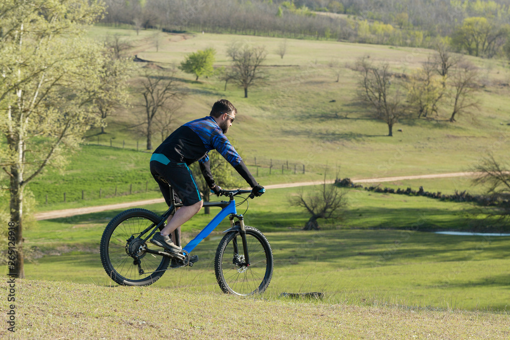 Fototapeta premium Cyclist in shorts and jersey on a modern carbon hardtail bike with an air suspension fork rides off-road on green hills near the forest