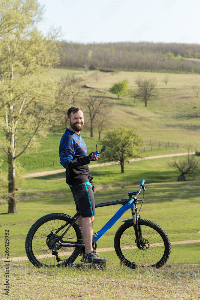 Obraz premium Cyclist in shorts and jersey on a modern carbon hardtail bike with an air suspension fork rides off-road on green hills near the forest