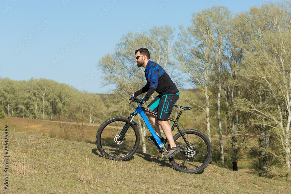 Cyclist in shorts and jersey on a modern carbon hardtail bike with an air suspension fork rides off-road on green hills near the forest Cyclist in shorts and jersey on a modern carbon hardtail bike with an air suspension fork rides off-road on green hills near the forest