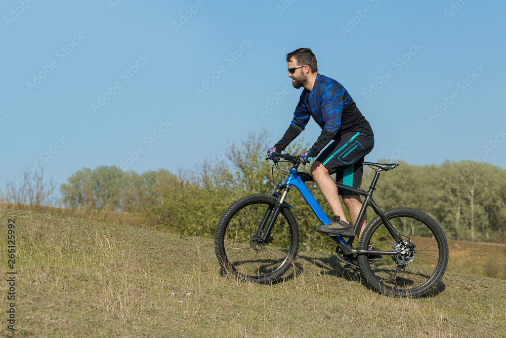 Cyclist in shorts and jersey on a modern carbon hardtail bike with an air suspension fork rides off-road on green hills near the forest