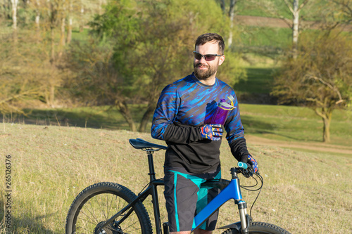 Cyclist in shorts and jersey on a modern carbon hardtail bike with an air suspension fork rides off-road on green hills near the forest