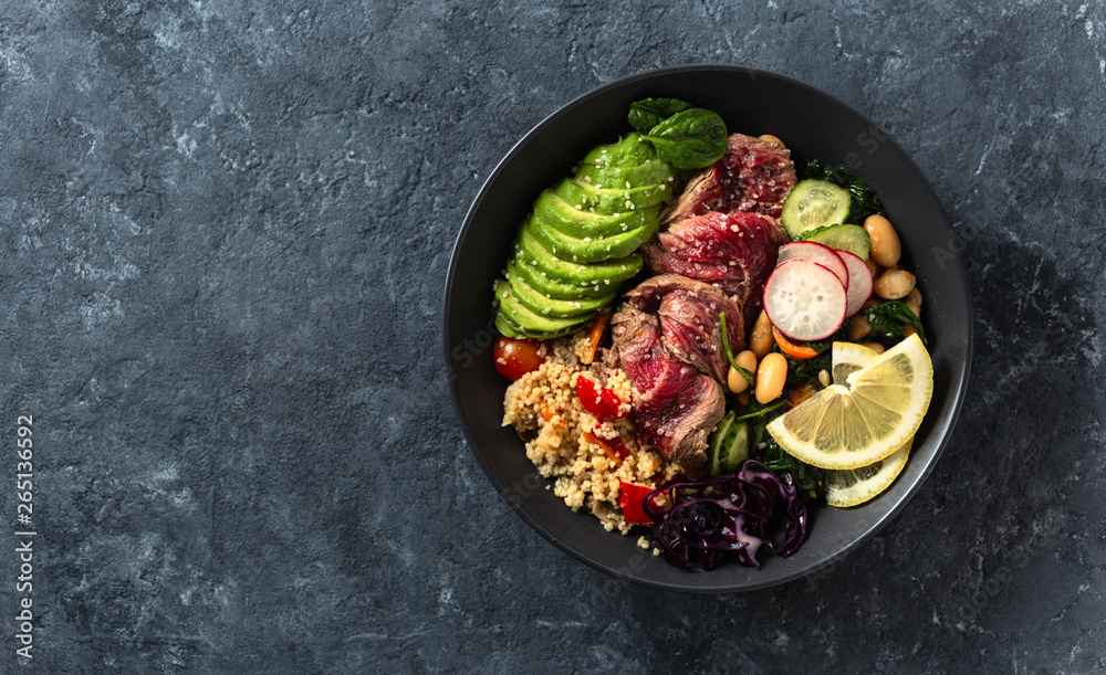 Healthy food buddha bowl with beef steak, beans, couscous, avocado and vegetables on dark background with copy space