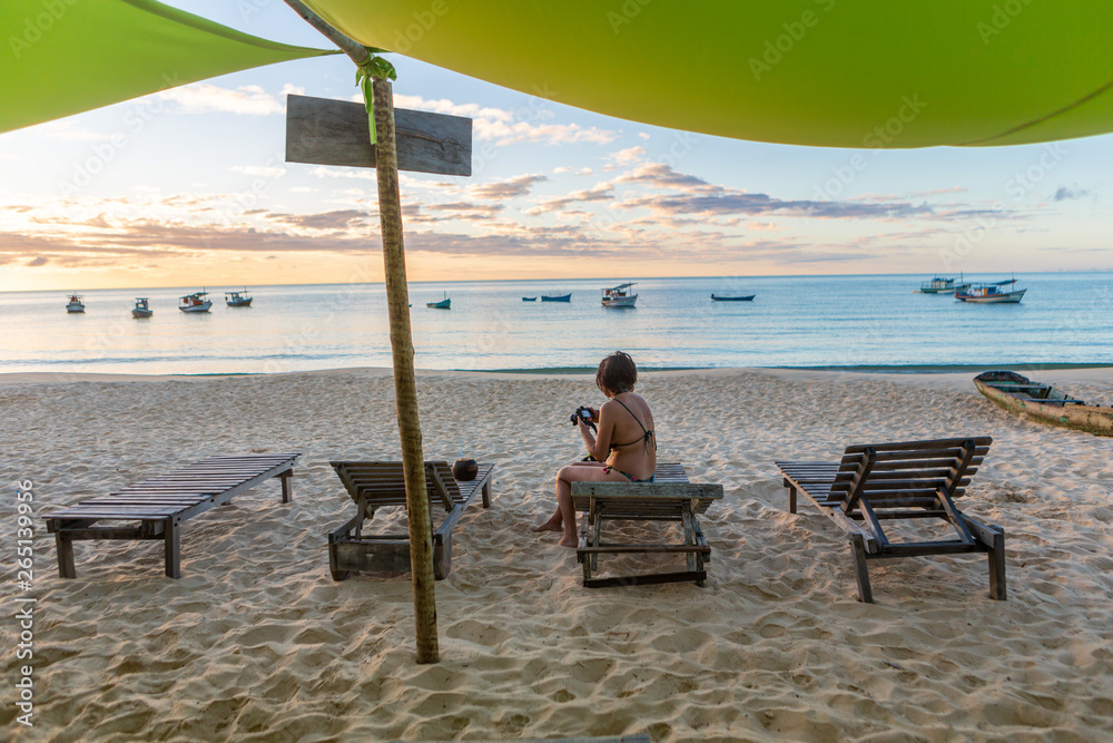 Beautiful young woman photographing sunset on idyllic beach sitting on chaise longue on sand with sea and boats in background. Conceito de férias, paz e relaxamento. Ponta do Corumbau, Bahia, Brasil.
