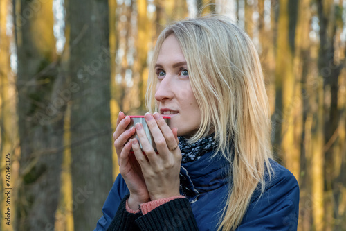 A young blonde girl drinks tea from a thermos cup in the spring forest at sunset. Theme travel. Girl drinking tea during a hike