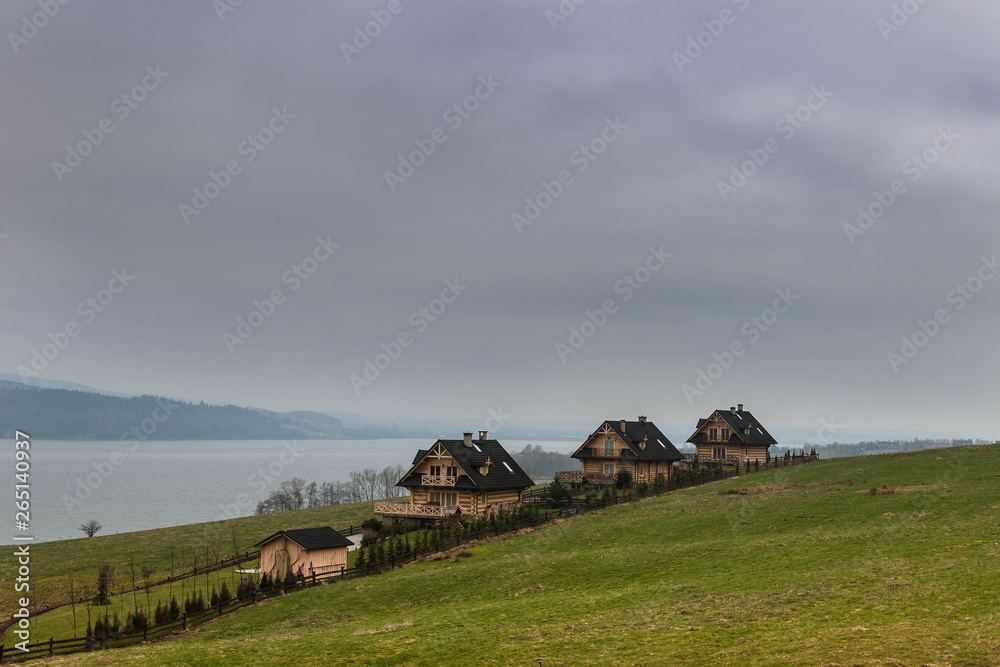 Foggy landscape with three wooden houses on the grass green hill at the shore of the lake on a cloudy day.