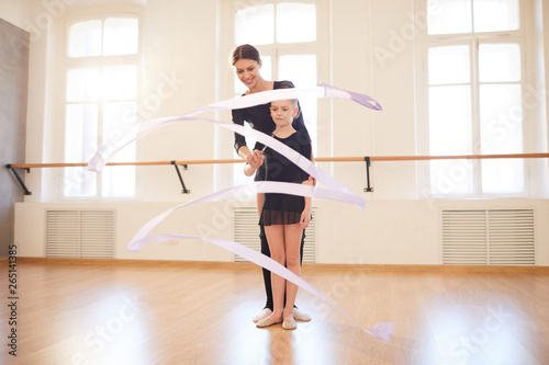Full length portrait of teacher helping little girl doing ballet moves in studio lit by sunlight, copy space