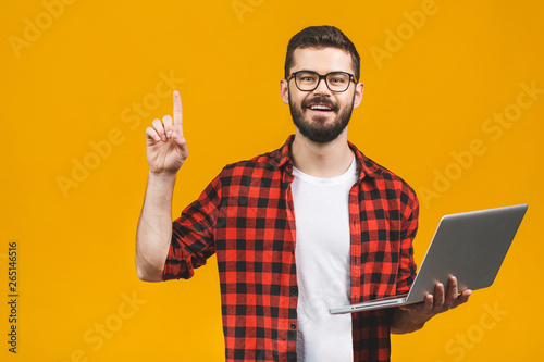 Young bearded man holds laptop and finger point up isolated against yellow background.