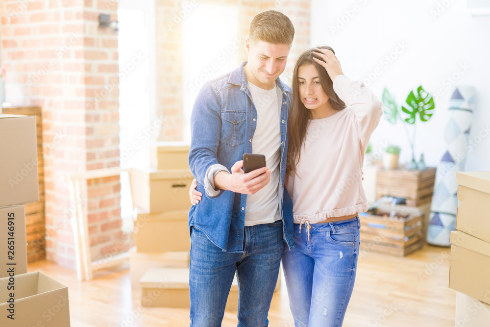 Beautiful young couple moving to a new house using smartphone stressed with hand on head, shocked with shame and surprise face, angry and frustrated. Fear and upset for mistake.