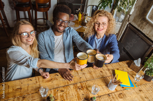 Top view of three multiethnic friends drinking latte at coffee bar restaurant. People in eyeglasses works  sits at wooden table clinking cups together at cafeteria. Friendship concept