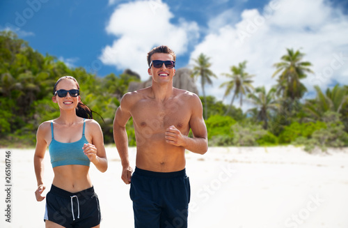 fitness, sport and lifestyle concept - happy couple in sports clothes and sunglasses running over tropical beach on island of seychelles background