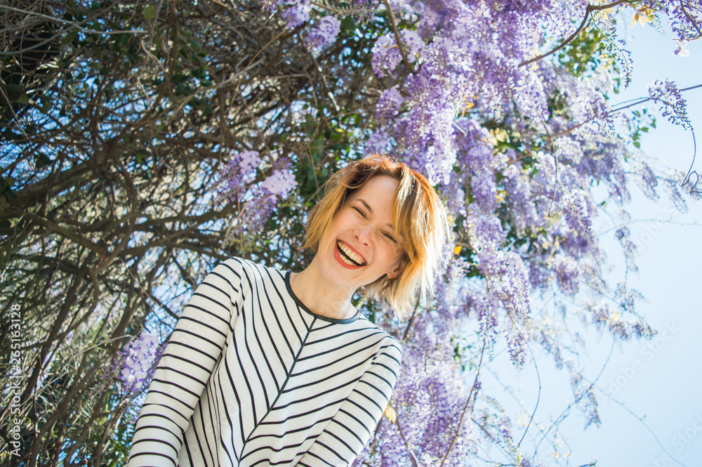 A beautiful woman with a short haircut is laughing and having fun in a blooming wisteria in the street with disheveled hair. A beautiful woman with a short haircut is laughing and having fun in a blooming wisteria in the street with disheveled hair.
