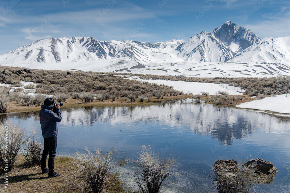 Asian man tourist and photographer holding DSLR camera taking photo of Mammoth mountain landscape from the lake in Hot Creek Geological Site in winter. Travel photography concept