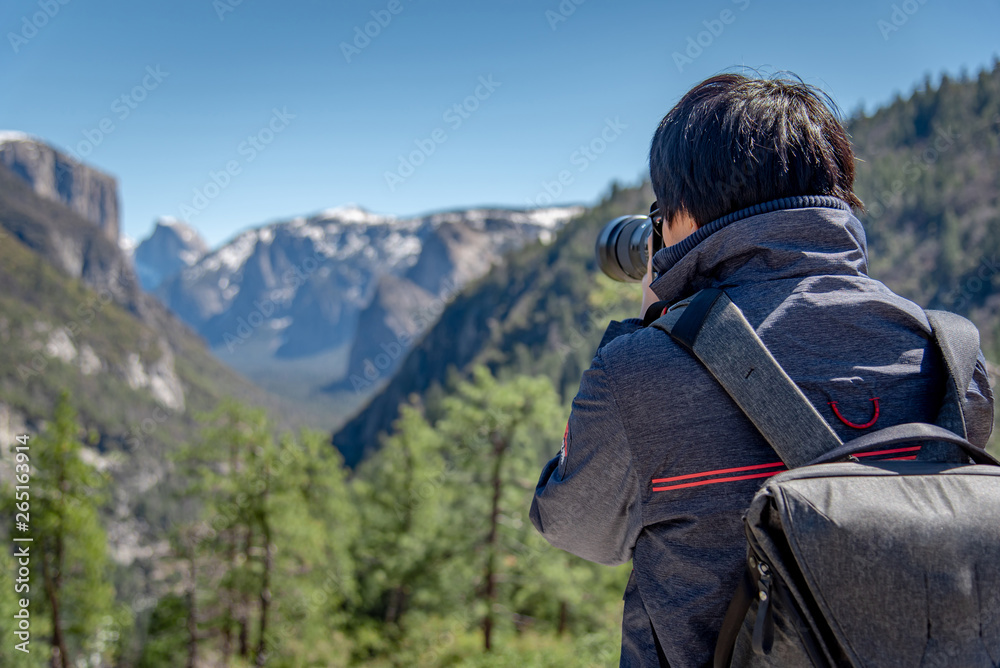 Asian man photographer and tourist with backpack holding DSLR camera taking photo of Half Dome mountain in Yosemite National Park, natural attraction in California, USA. Travel photography concept