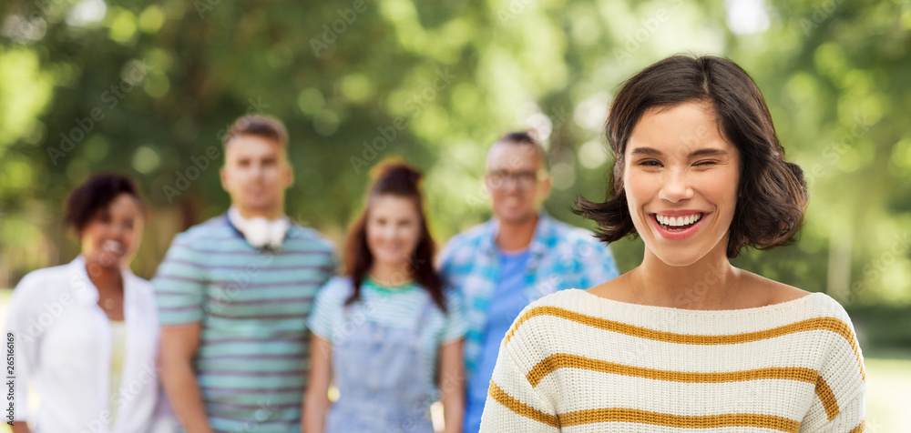 expression and people concept - happy smiling young woman in striped pullover winking and group of friends in summer park on background expression and people concept - happy smiling young woman in striped pullover winking and group of friends in summer park on background