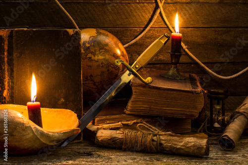 Old books and documents, a burning candle, globe and dirk stand on a wooden surface.