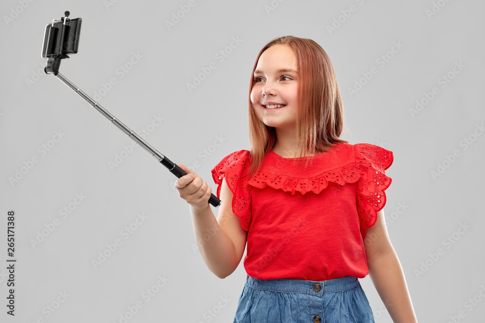 childhood and people concept - beautiful smiling girl in red shirt and skirt taking picture by smartphone on selfie stick over grey background childhood and people concept - beautiful smiling girl in red shirt and skirt taking picture by smartphone on selfie stick over grey background