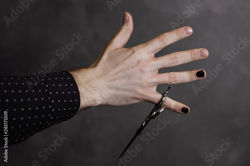 The male hand with manicure holds scissors for a hairstyle on a gray background. Nails are painted black color.