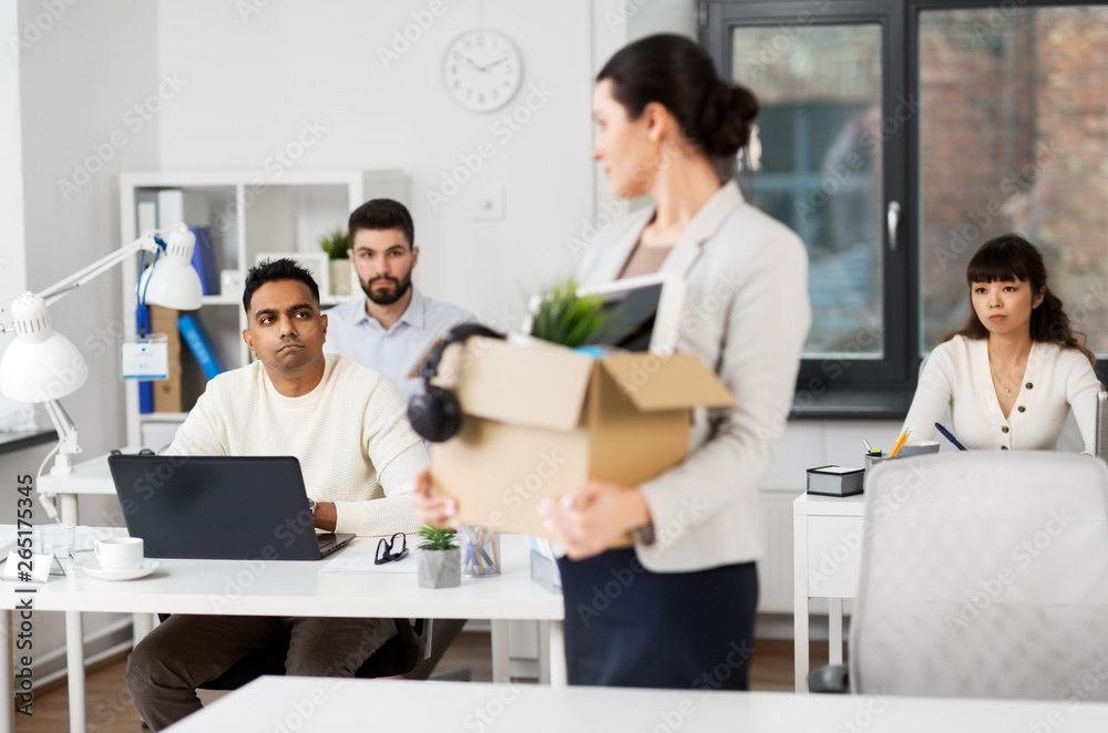 business, firing and job loss concept - sad fired female employee with box of personal stuff leaving office looking at her colleagues