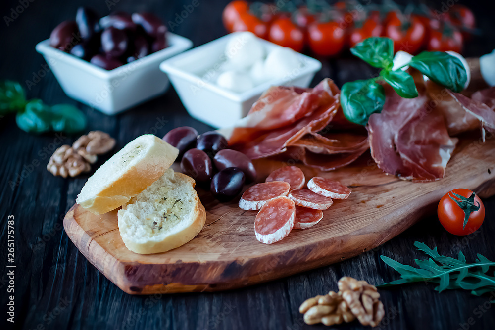 Prosciutto, bread, olives, walnut, mozzarella, salami, basil and cherry tomatoes on brown wooden board. Mediterranean kitchen. Prosciutto, bread, olives, walnut, mozzarella, salami, basil and cherry tomatoes on brown wooden board. Mediterranean kitchen.