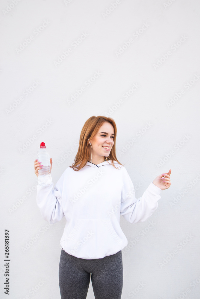 Fototapeta premium Happy, attractive girl on a white background, holding a bottle of water in her hands and listening to music in the headphones and looking away. Portrait of joyful fitness girl.
