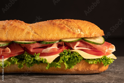 Huge fresh crispy baguette sandwich with meat, prosciutto, cheese, lettuce salad and vegetables. Close up. Black background. Space for text.