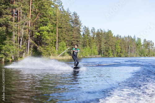 Young pretty slim brunette woman in wetsuit riding wakeboard wave of motorboat