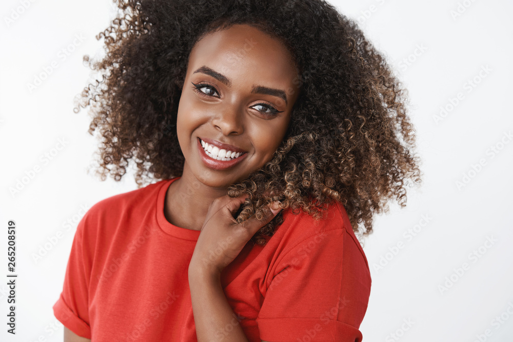Close-up shot of carefree happy and gentle gorgeous african-american female model with curls smiling broadly touching hair, gazing delighted, tender at camera wearing red t-shirt over white background Close-up shot of carefree happy and gentle gorgeous african-american female model with curls smiling broadly touching hair, gazing delighted, tender at camera wearing red t-shirt over white background