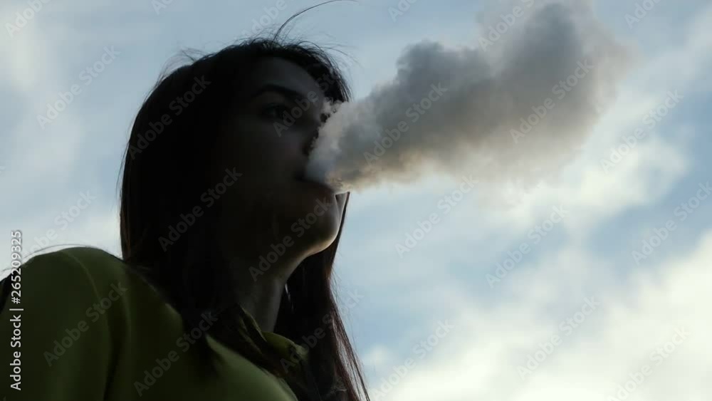Dreamy caucasian girl vaping at sunset outdoors. Silhouette smoking woman on the cloudy sky background in Slow motion Dreamy caucasian girl vaping at sunset outdoors. Silhouette smoking woman on the cloudy sky background in Slow motion