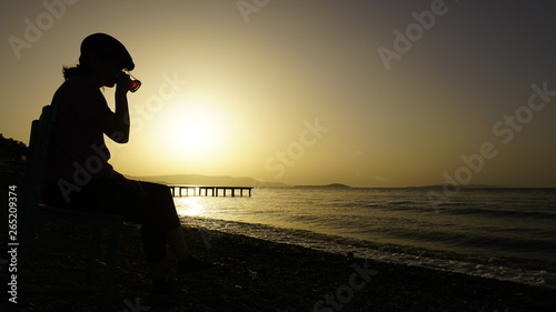 Young woman drinking tea or coffee, watching sea on chair on pebble beach towards dramatic sunset. Amazing sunset over mountains & ocean horizon at weekend. Orange and yellow sunny day at outdoor cafe