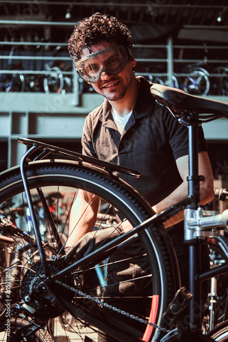 Smiling diligent master in protective glasses is fixing bicycle at workshop.