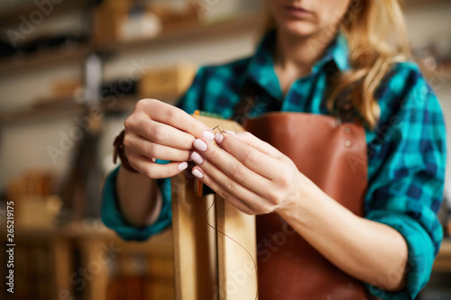 Close up of young woman holding thread and needle while making bags and shoes in leatherworking shop, copy space