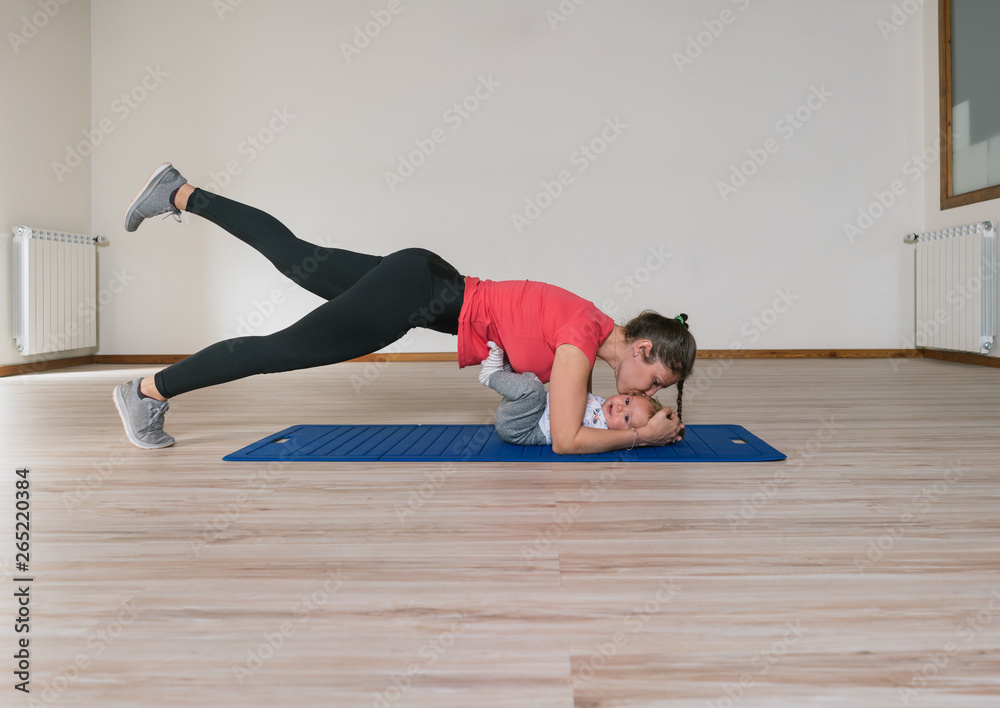 Side view of woman in sportswear kissing baby and exercising on floor during fitness training in gym Side view of woman in sportswear kissing baby and exercising on floor during fitness training in gym