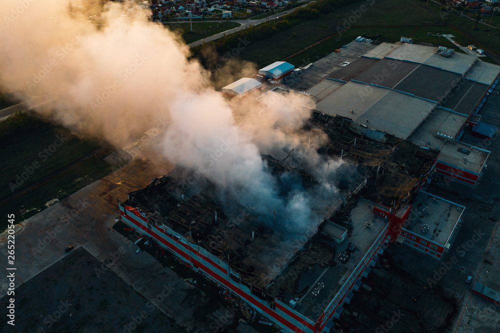 Fototapeta premium Aerial view of burnt industrial warehouse or logistics center building after big fire with huge smoke from burned roof, drone shot