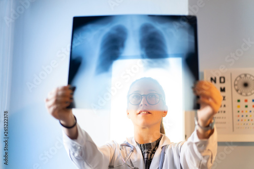 Front view of young female doctor in uniform looking at x-ray image on wall in room