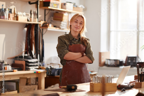 Warm toned portrait of confident female artisan standing with arms crossed while posing in workshop photo