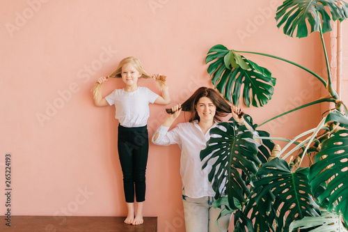 Life style female family portrait of happy mother and smiling daughter make same poses on pink wall with palm tree branches home background. Cheerful mom with her blonde daughter posing identically. photo