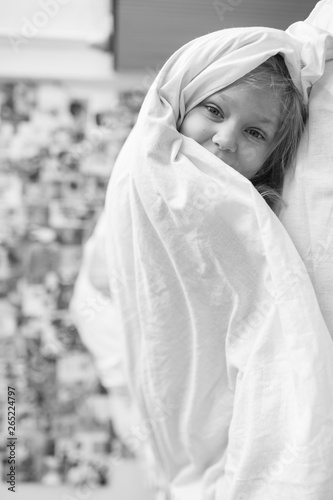 Vetrtically turned black white portrait of little girl lying on bed at home under white blanket and looking ar camera with decorative wall on background.