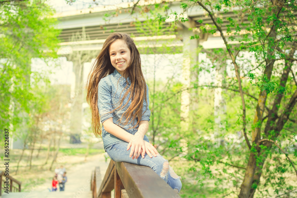 Glad girl sitting on railing Pleased girl in casual outfit looking at camera while sitting on wooden railing of stairway in green park Glad girl sitting on railing Pleased girl in casual outfit looking at camera while sitting on wooden railing of stairway in green park