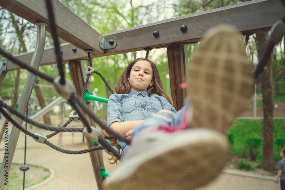 Obraz premium Cute girl having fan on playground. Charming little brunette girl in jeans suit hanging on grid and looking at camera on playground in park