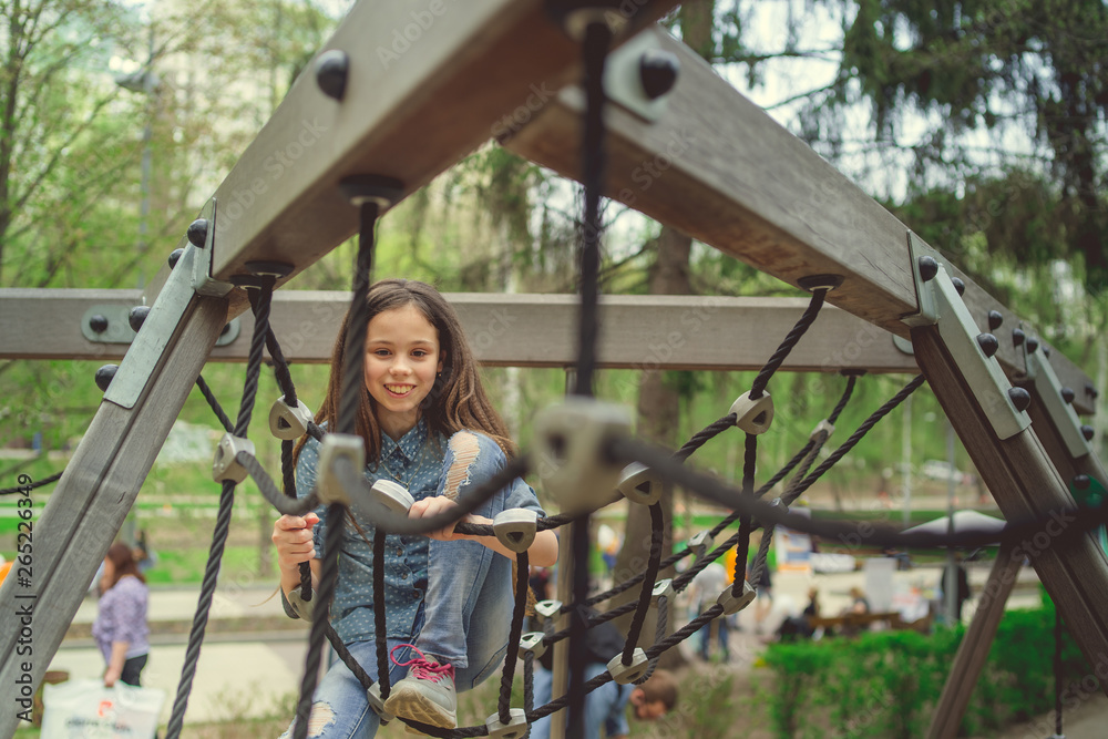 Obraz premium Cute girl having fan on playground. Charming little brunette girl in jeans suit hanging on grid and looking at camera on playground in park