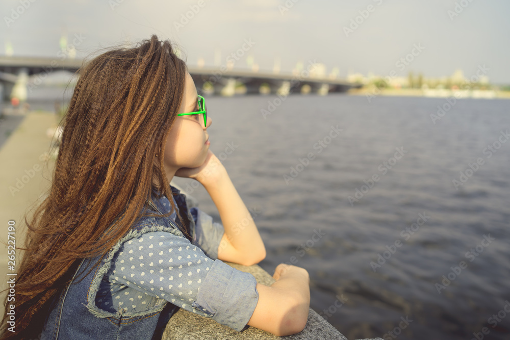 Girl in sunglasses near river Portrait of adorable little brunette girl in sunglasses and denim suit sitting on column near river on sunny day