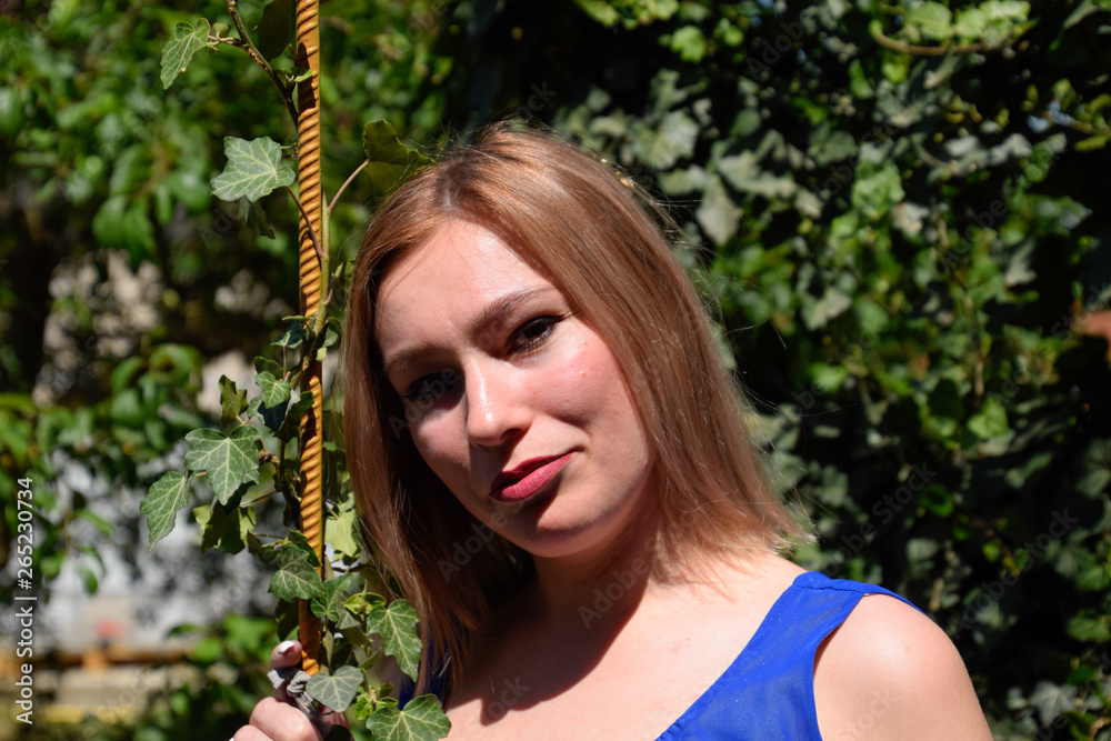 Naklejka premium Portrait of a girl in a blue dress on a background of green arbor of ivy.