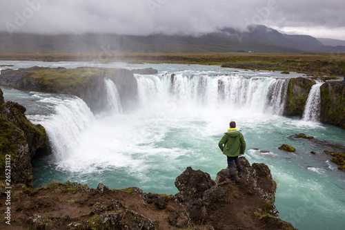 A male tourist stands on a cliff above a waterfall and admires its beauty. Picture taken in Iceland.