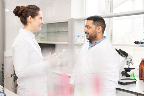 Positive confident multi-ethnic laboratory workers in white coats standing in scientific laboratory and talking about research plan