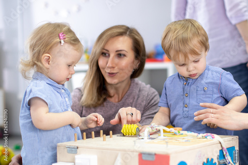 Cute toddlers babies playing with busyboard. Mother or caregiver teaching to kids in nursery. Children's educational toys.