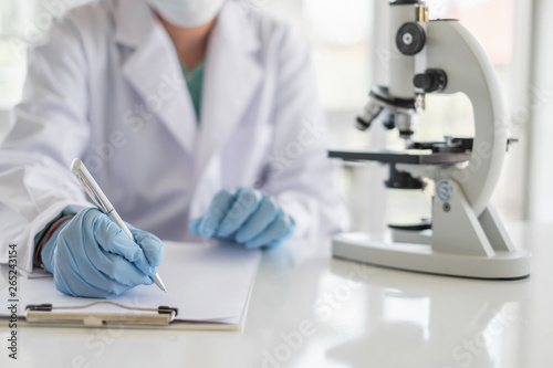 A scientist hands writing on a clipboard in laboratory with test tube microscope and solutions.