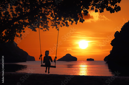 Silhouette of a girl sitting on a swing or cradle on the beach at sunset. © THANAGON