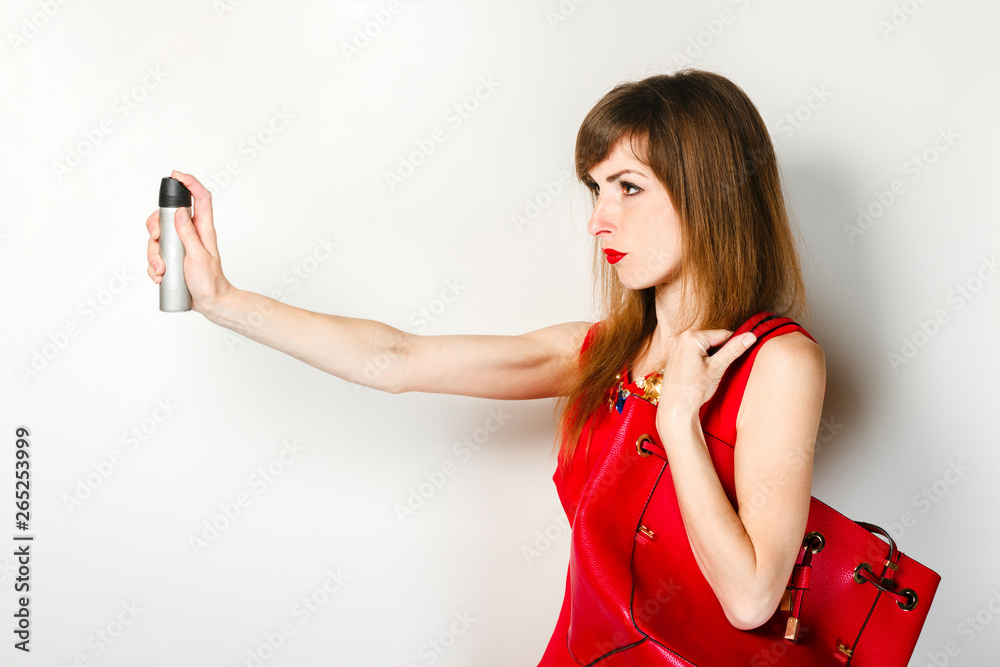 Naklejka premium Young girl in a red dress holds a gas spray in her hand for protection against a light background. Concept of an attack on a girl, protection from a burglar, gangster.